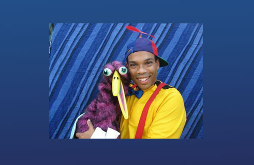 Tommy Terrific is wearing a yellow buttoned down-shirt with wide red suspenders, a cap with a plastic propeller on top while holding a larger purple bird puppet.  