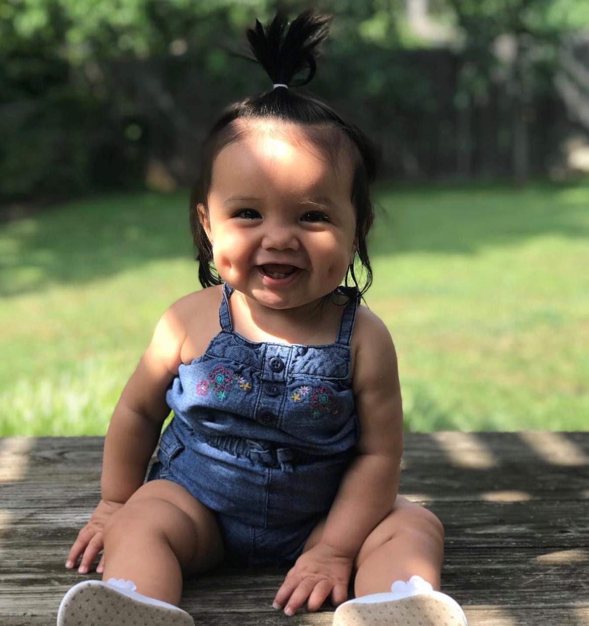 Smiling baby girl in overalls sitting on a picnic table under the shade of a tree.