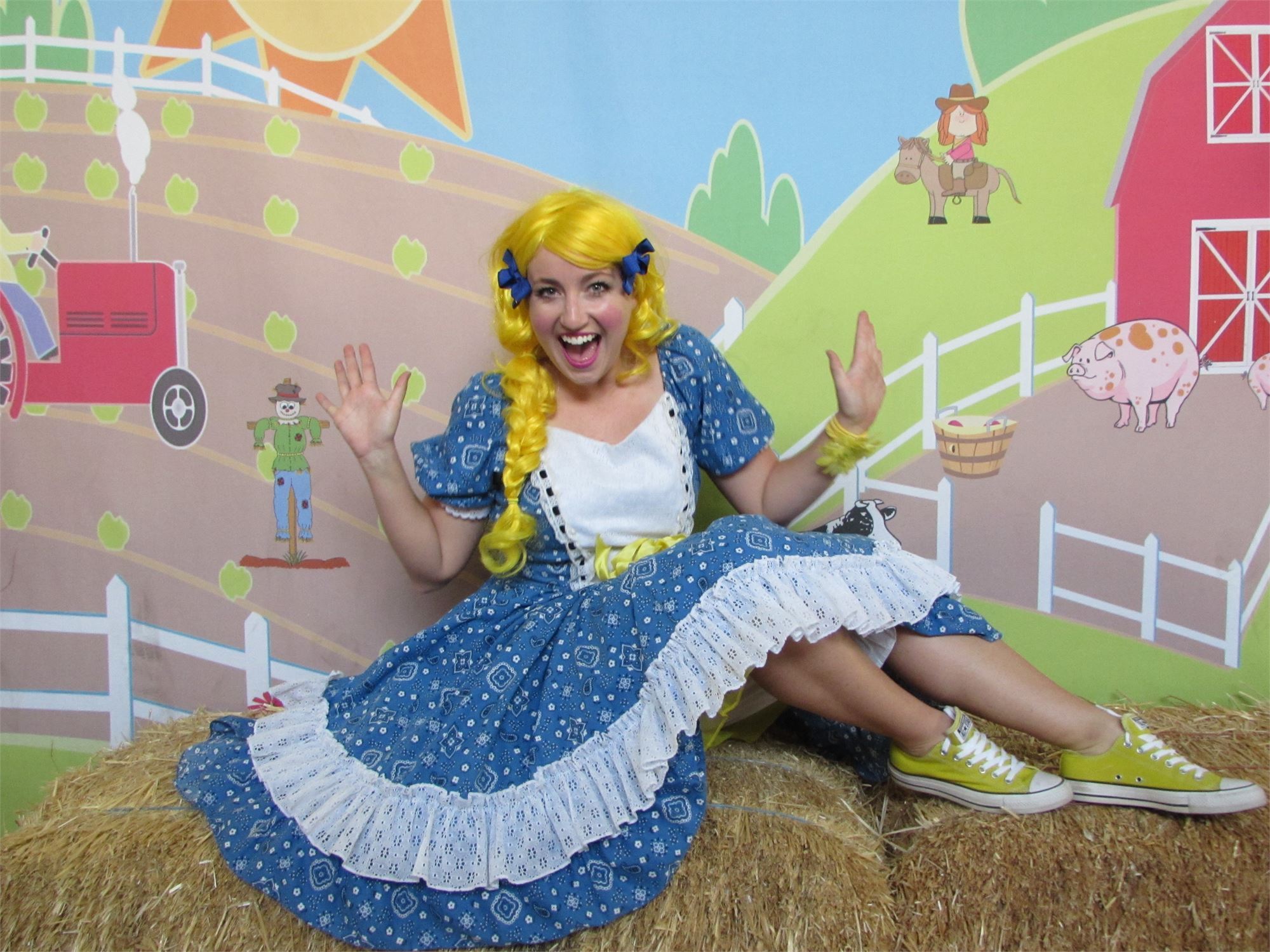 A young woman wearing a long yellow wig and a blue and white dress sitting on a bale of hay.