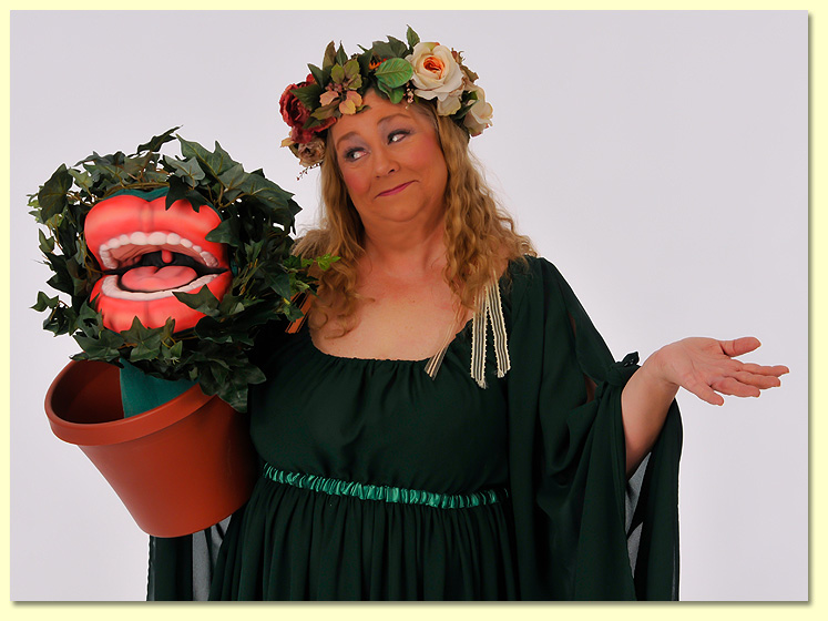 A woman with long blonde hair wearing a green a dark green dress and flowers in her hair is holding a Venus Flytrap puppet while shrugging happily.