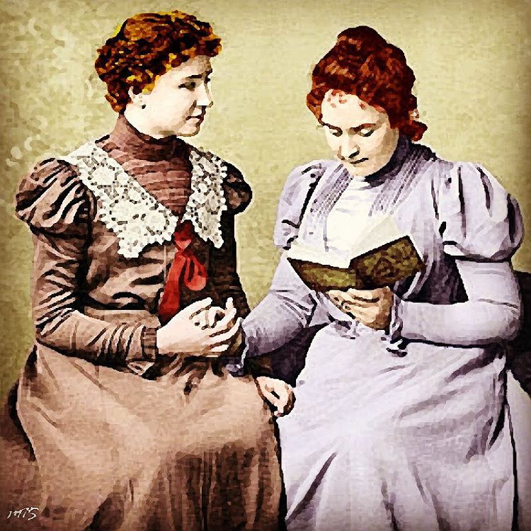 Two women sitting together holding hands while one is holding a book. One of the women is Helen Keller and the other is Anne Sullivan