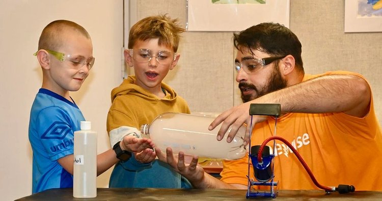 Photo of two elementary school children and an adult, all in safety goggles. They are pouring bubbles from a 2 liter bottle into the children's hands. All three are grinning. 