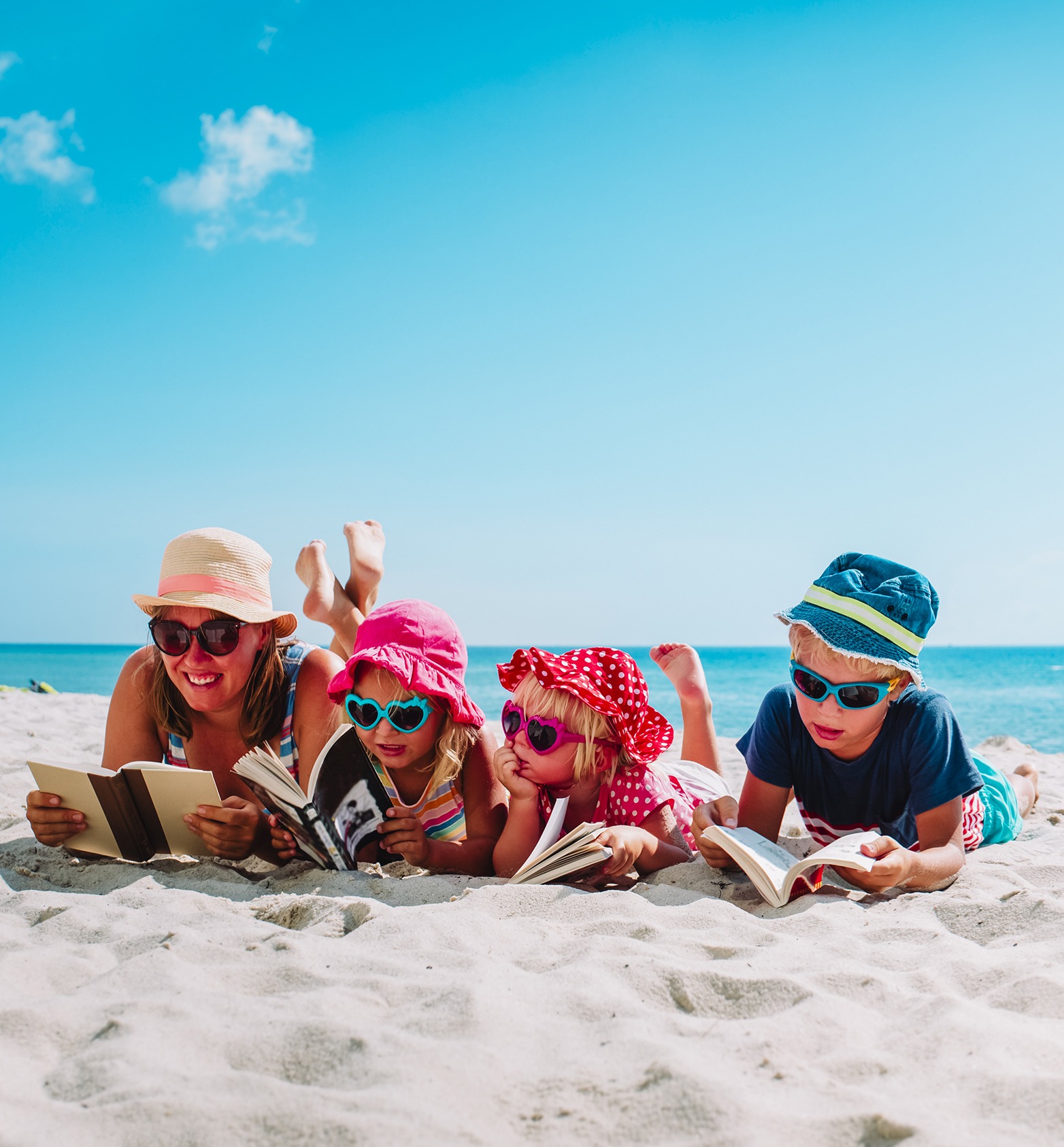 A woman and four children in sunglasses laying down on the beach, reading books.