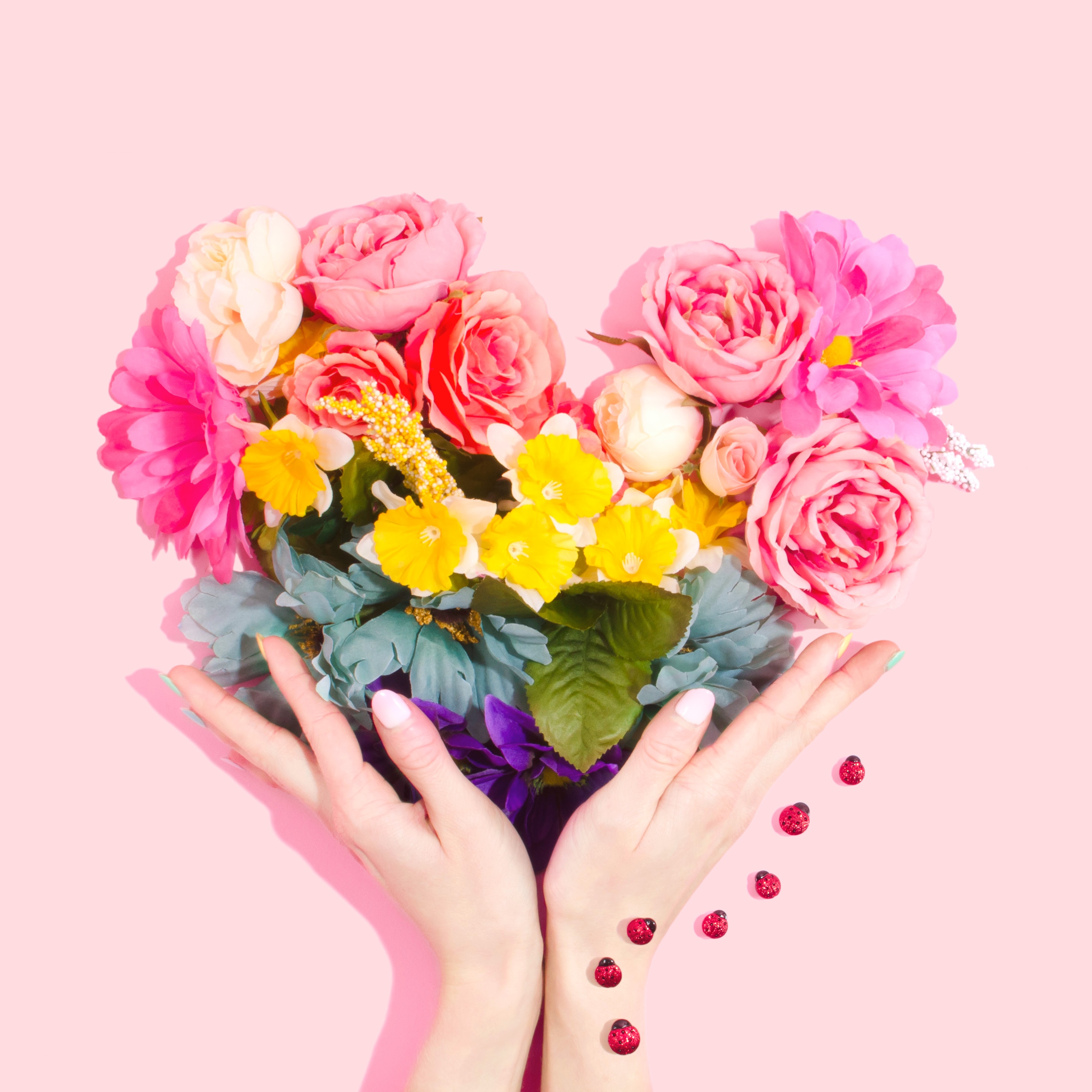 On a pink background, two hands offer up a heart made up of many different flowers.