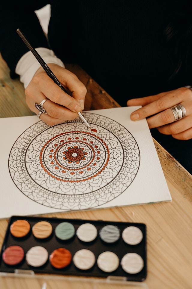 A person sits at a table with a paint brush in their hand working on a colorful mandala painting on a white sheet of paper. A tray of paints sits on the table in front of them.