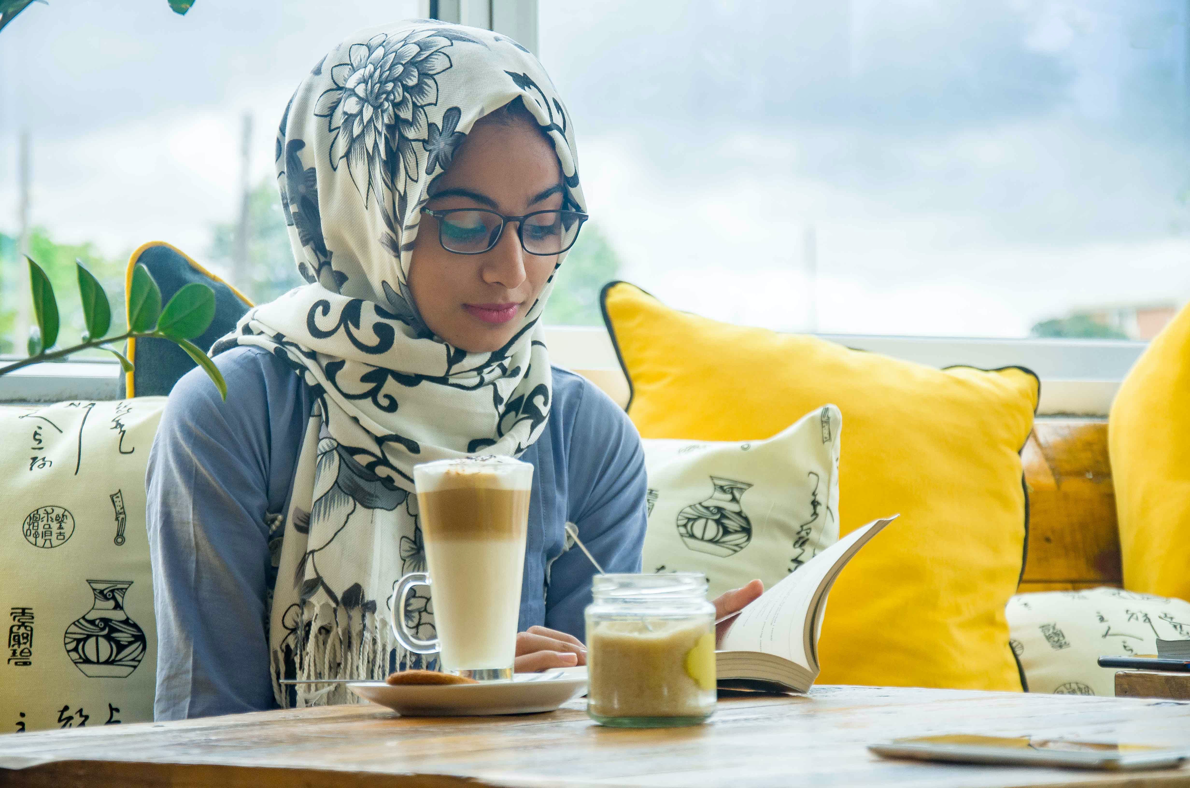 A woman with glasses wearing a head scarf reads with a coffee drink on the table in front of her.