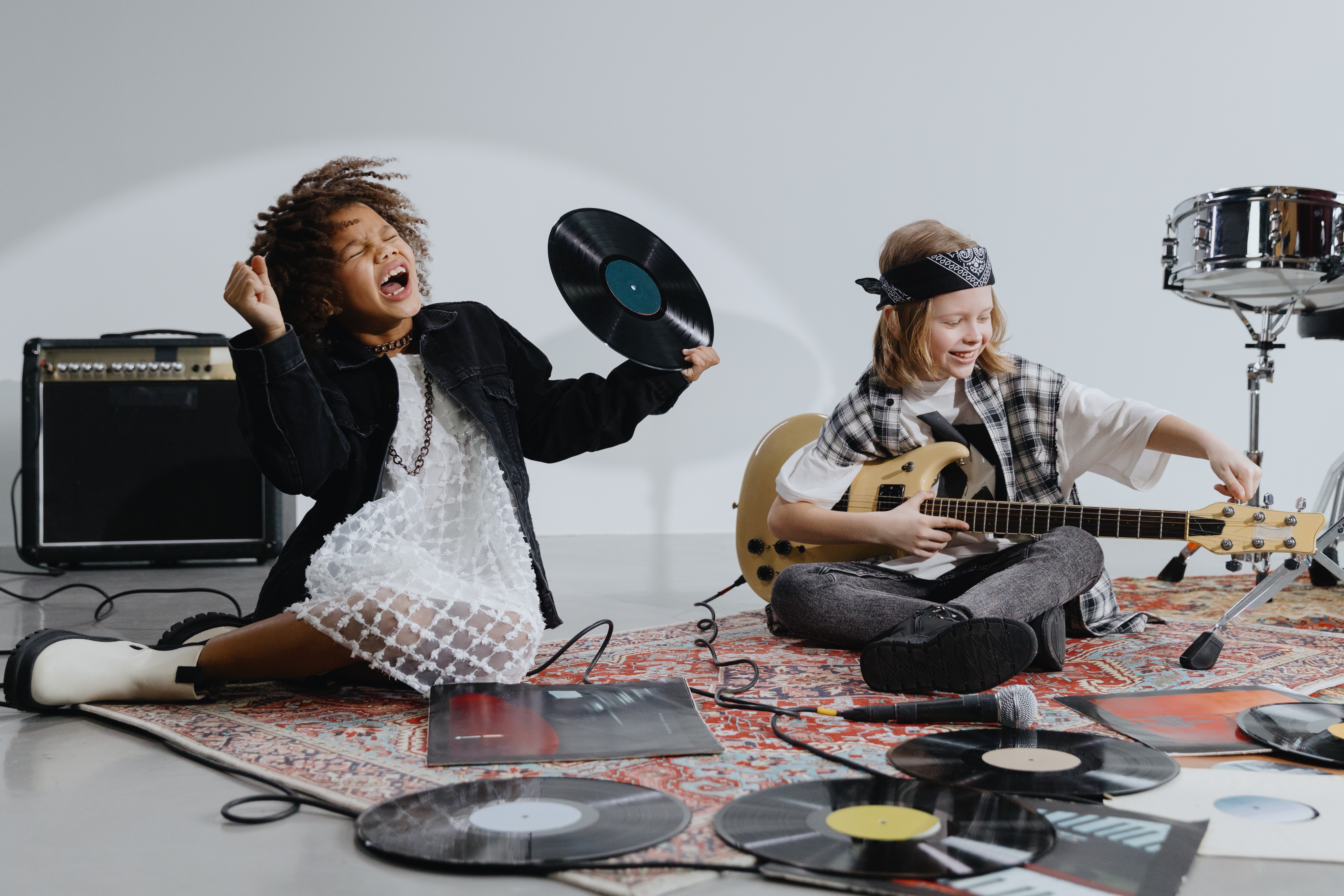 A young Black girl and young white boy sit on a rug surrounded by vinyl records. The boy is holding a guitar and the girl is holding a record and singing with her eyes squeezed shut.