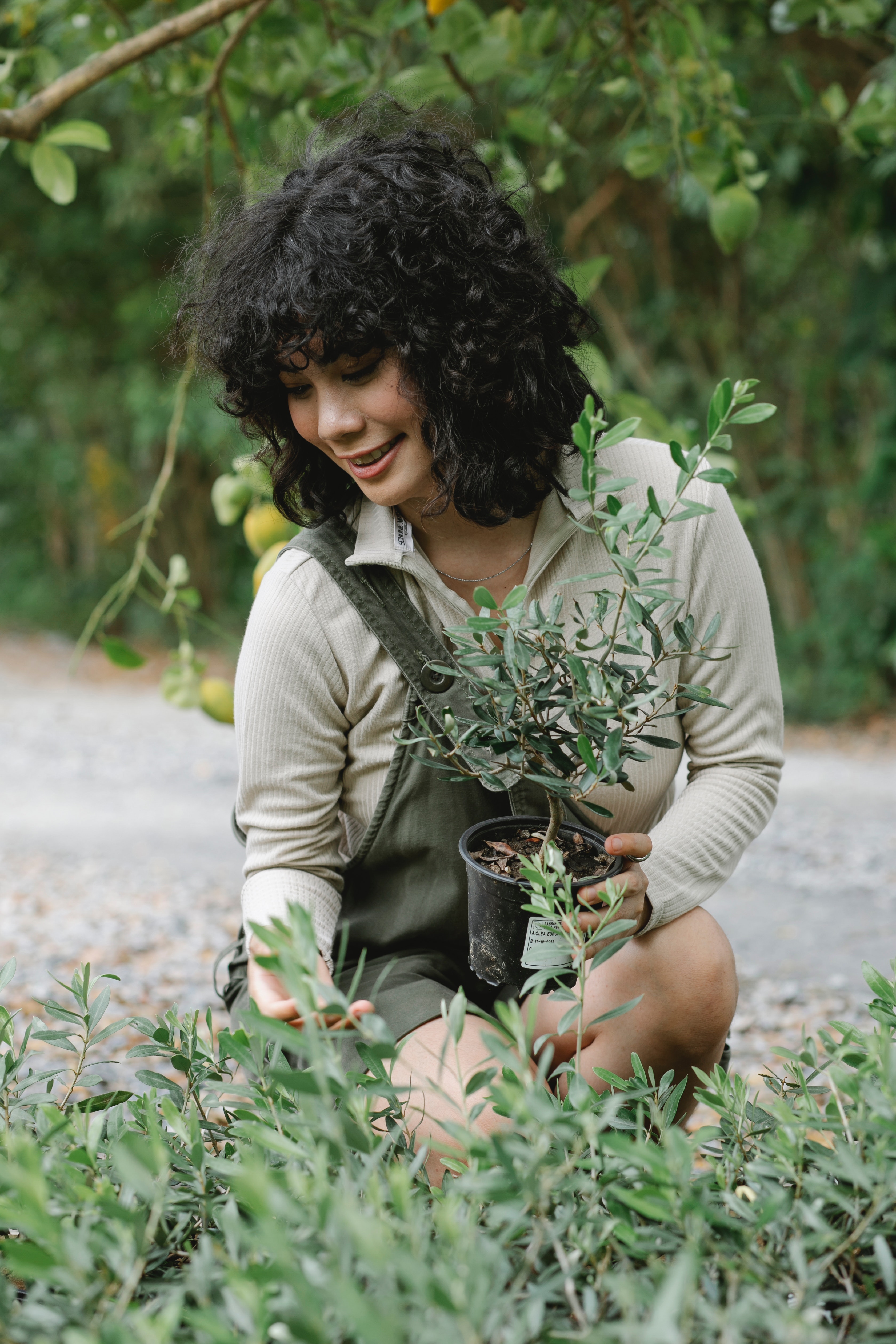 A young woman with dark, curly hair squats in front of a patch of garden holding a potted plant in her hand.