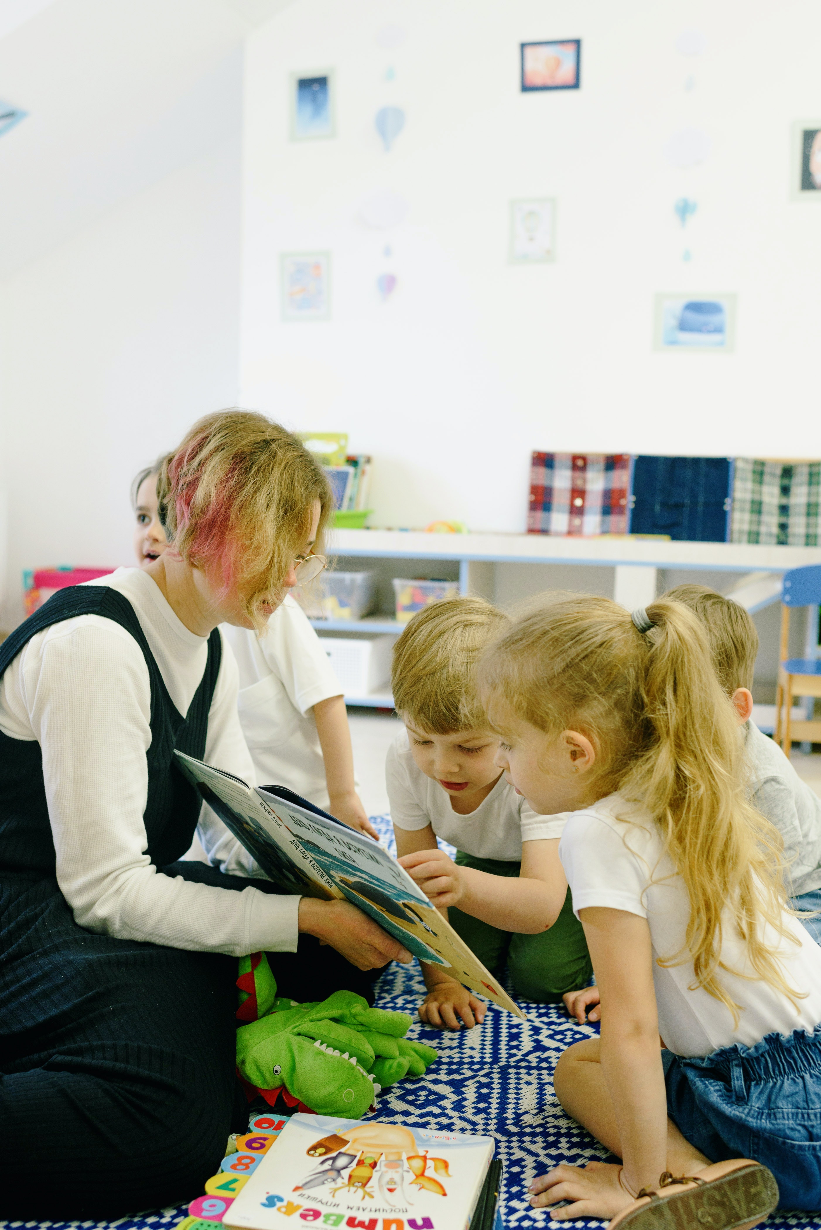 A blonde woman with a pink streak in her hair holds out a book to a group of children who are pointing and engaging with the text.