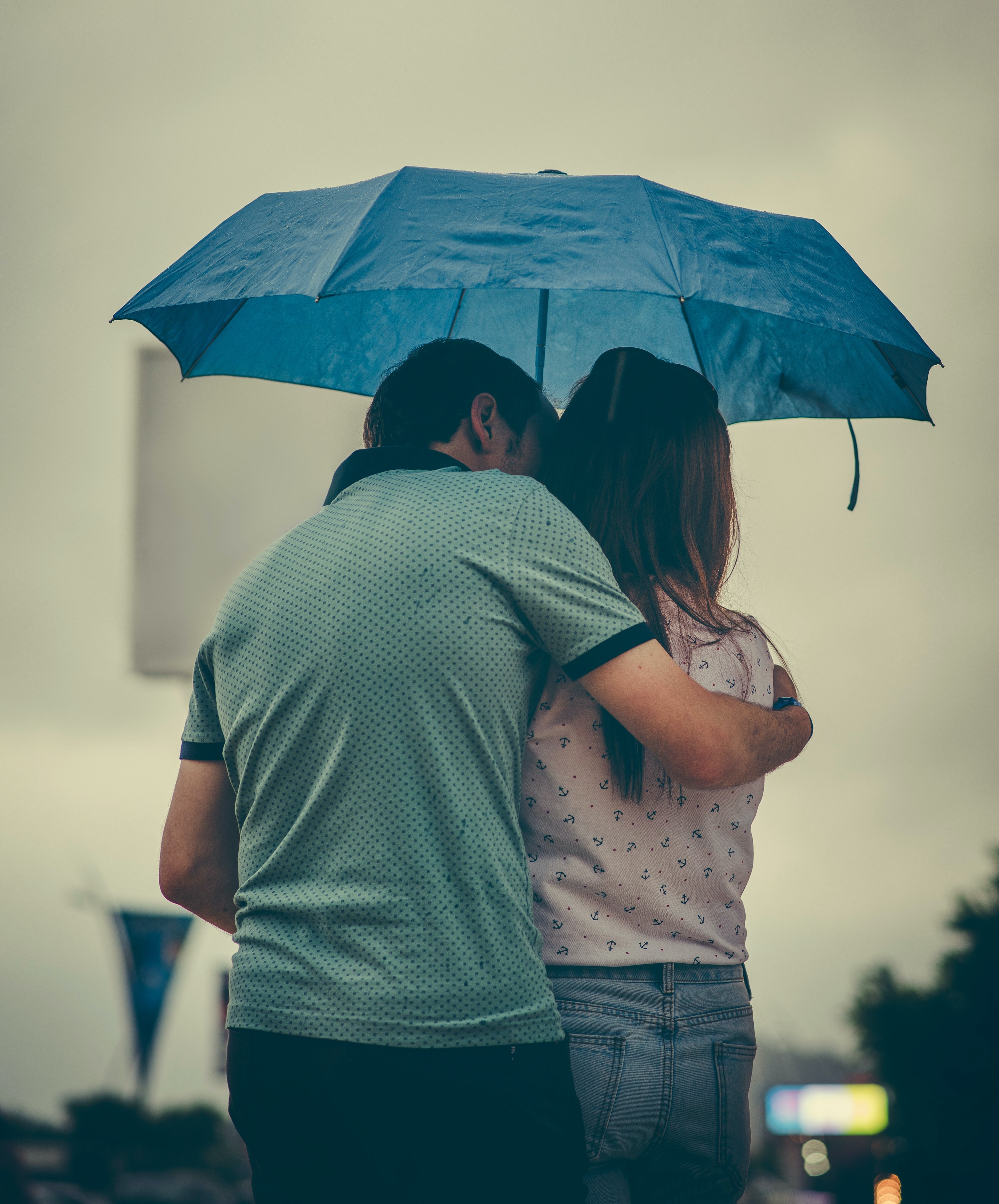 A couple under an umbrella with their backs to the camera, one person with their arm around the other as if to comfort them.