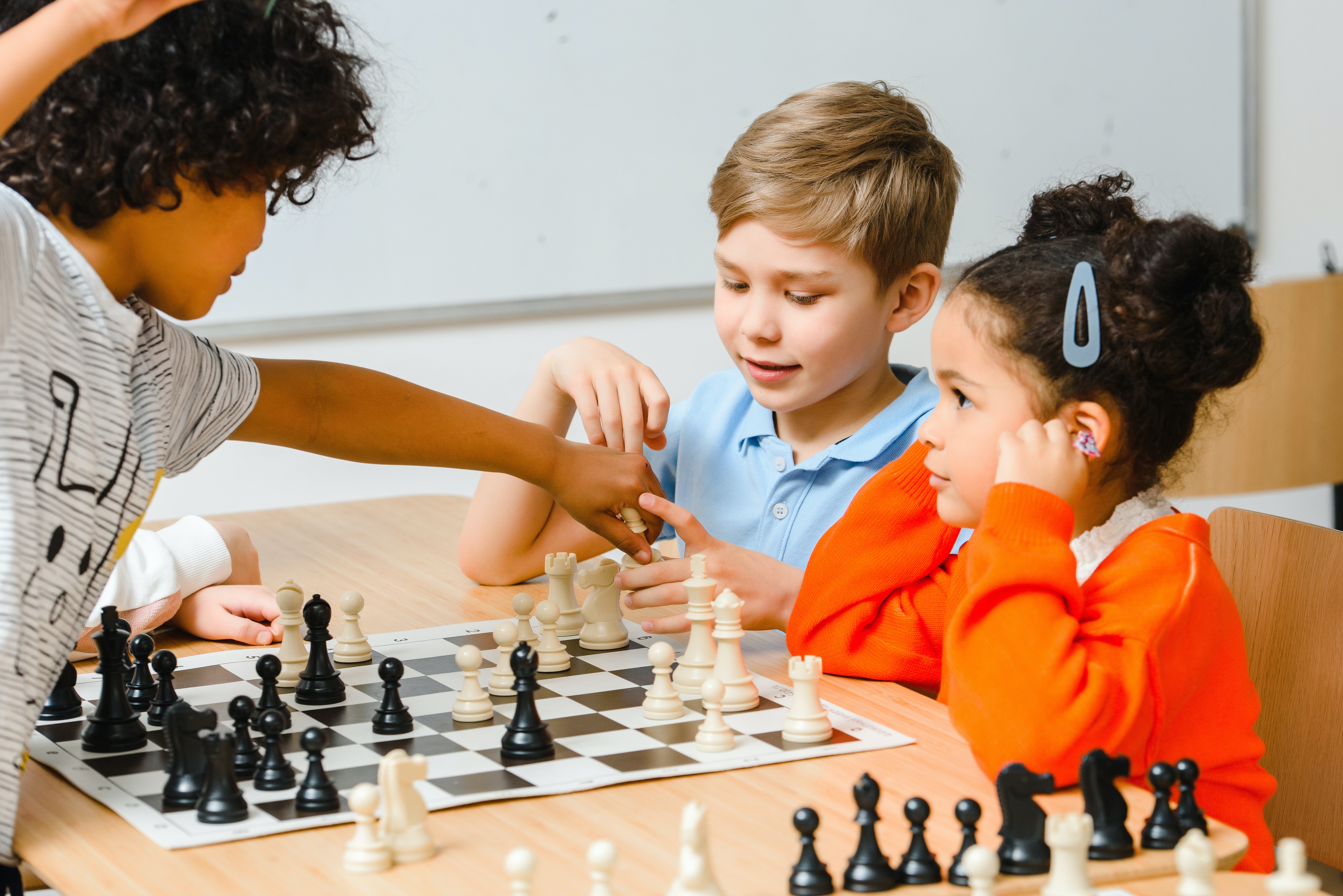 A group of kids are playing chess, one standing with his arm across the board reaching for a piece.