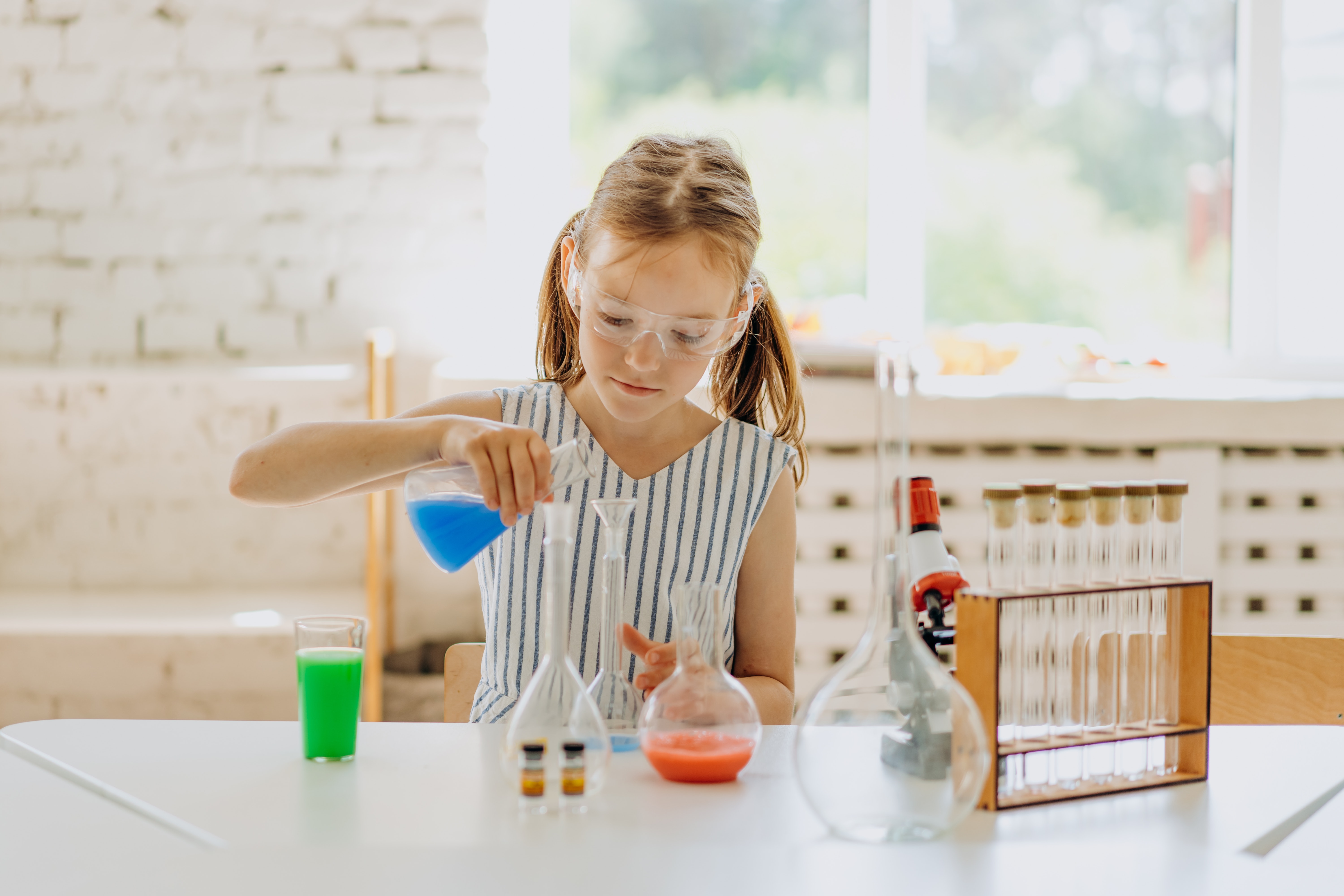 A young girl pours blue liquid from a glass beaker into another beaker on a table filled with tubes and beakers.