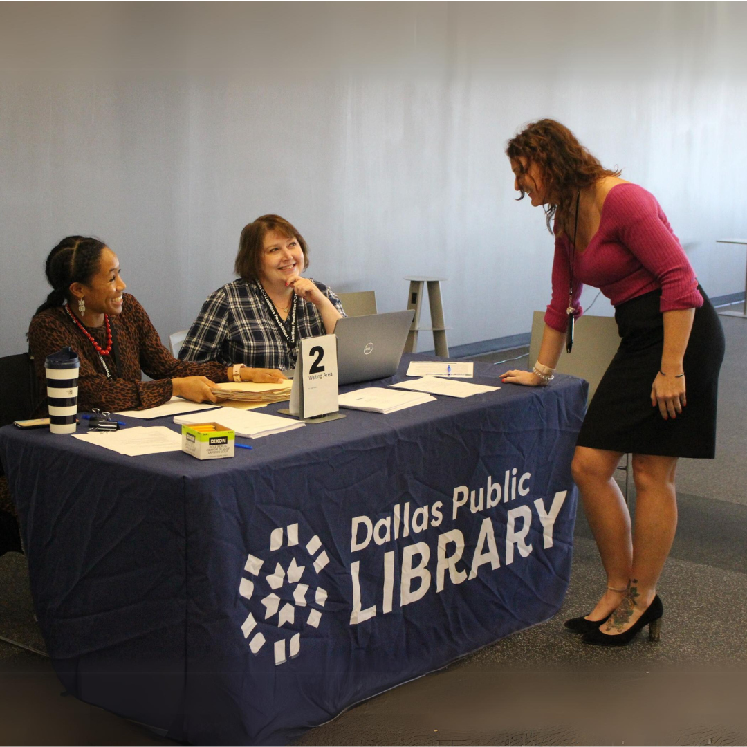 Two people sitting at a Dallas Public Library table and a woman leans over to talk to them