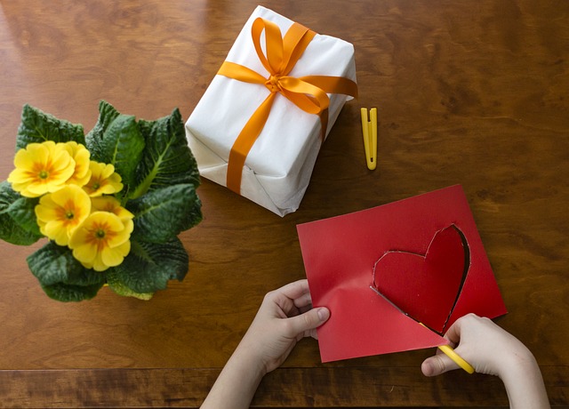 Child Hands Cutting out a Heart on Red Paper Next to a Gift Box and Yellow Flowers
