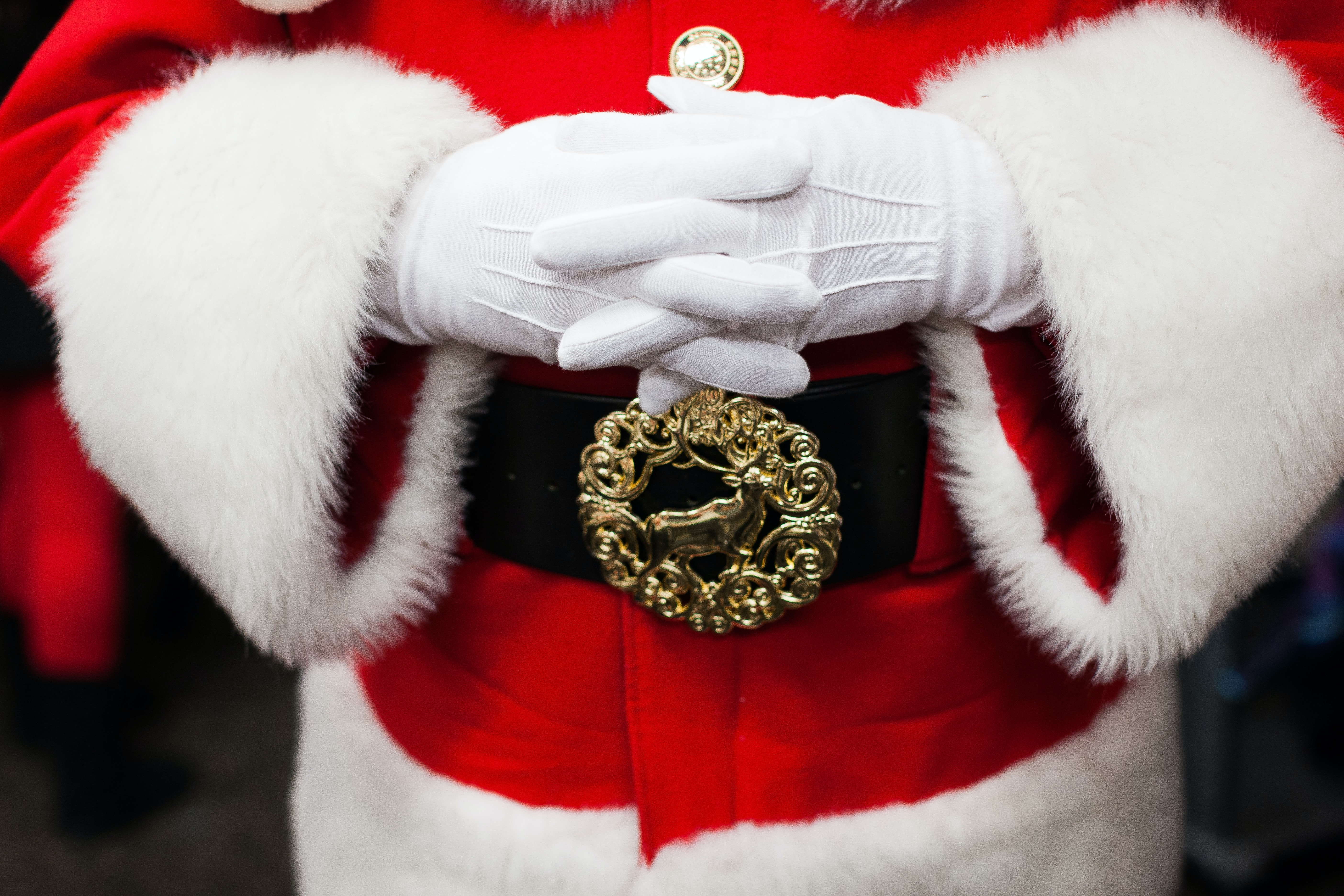 Santa in his traditional red and white suit, seen only from his chest to the bottom of his coat. His gloved hands are folded just above his big, black belt with a golden buckle featuring a reindeer inside a wreath.