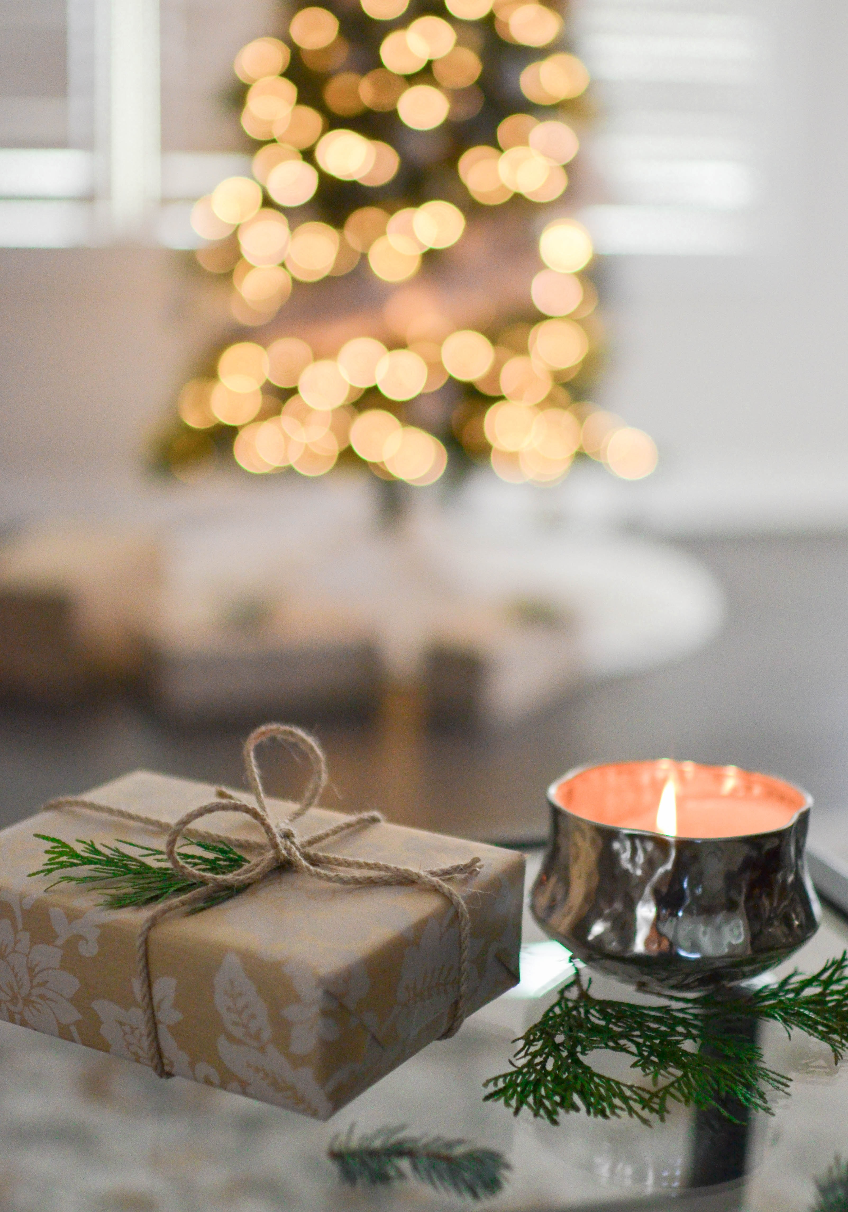 In the foreground, a wrapped gift tied with cord and decorated with a sprig of pine. Next to it, a candle in a small silver container burns with a pink glow. In the background is a lit and decorated tree with more gifts underneath it.