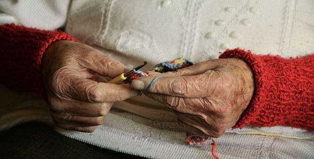 A close up of an older woman’s hands crocheting multicolored yarn. The woman’s sleeves are red crocheted fabric, and her torso is covered by a white sweater. 
