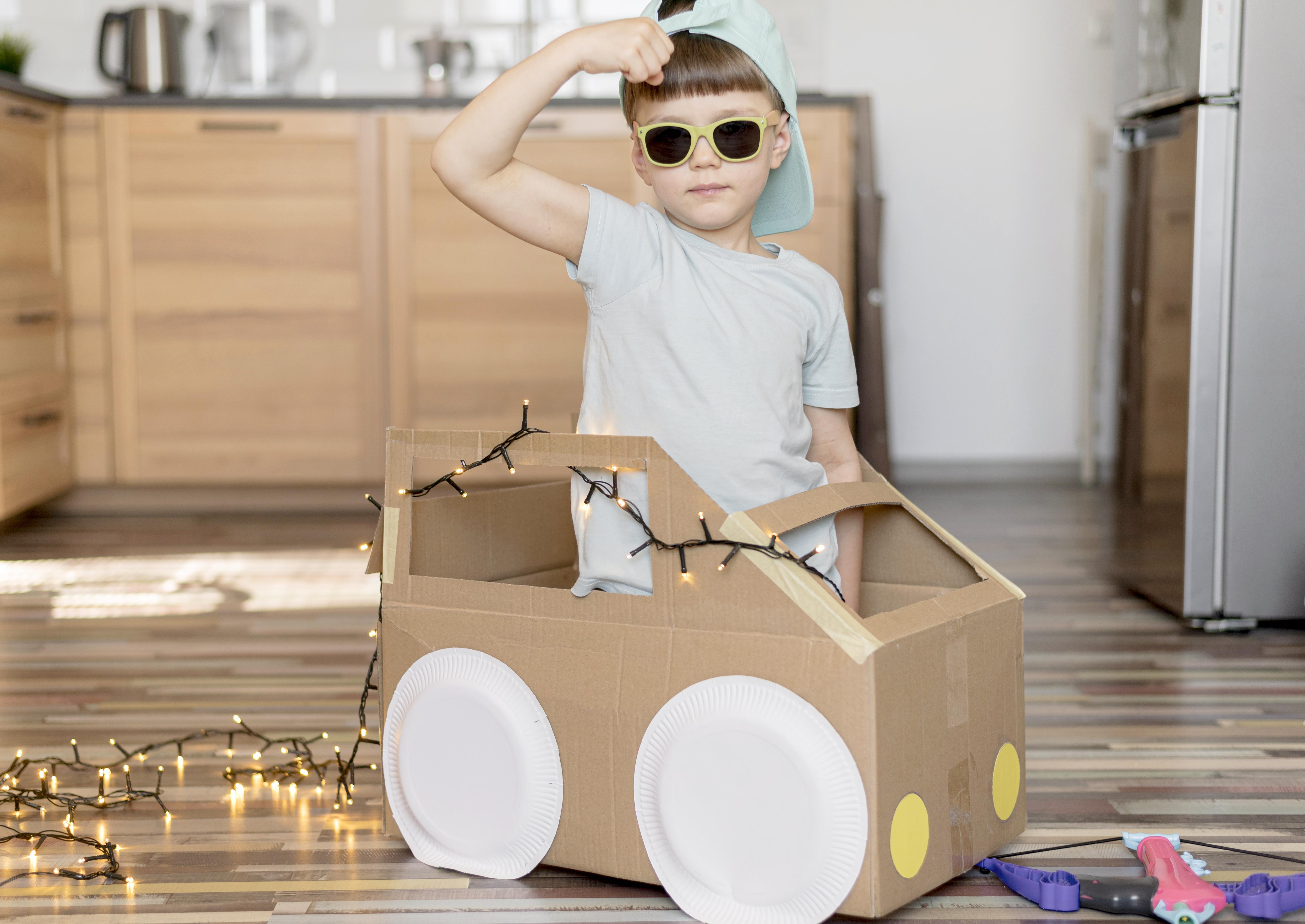 A young boy, looking cool, sitting inside a cardboard box car. The cardboard box car and cabinets behind it are brown. The boy is wearing a gray shirt, green framed sunglasses, and a pale blue hat. There are white Christmas lights coming out of the cardboard box car and trailing on the ground.
