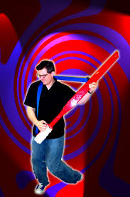 A man wearing glasses holding a giant toothbrush as he is pretending that it is a guitar in the foreground. In the background you see a psychedelic red and purple swirl. 