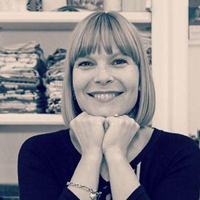 Woman (Regina Frydman) with a dark blonde bob haircut and bangs, smiling broadly with her chin resting on her hands. Shelves of craft materials in the background.