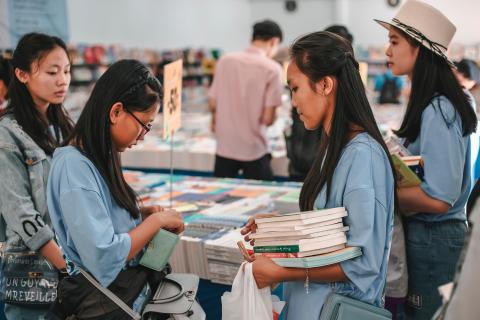 A group of women shop from a table stacked with books, one is holding a shopping bag and an armful of books