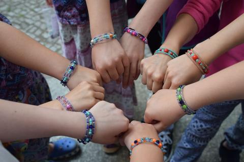 A group of young kids wearing beaded bracelets have their arms extended in a circle