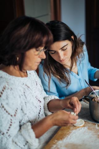 Two women work together on a cooking project, the younger woman leaning over to observe the older woman.