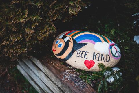 A rock painted with a bee and a rainbow with the text Bee Kind.