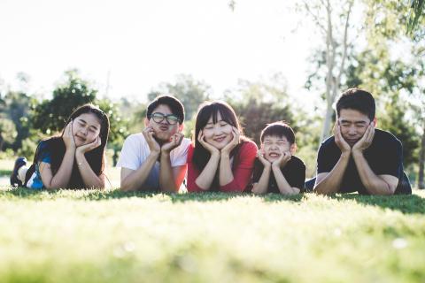 A family of five lays in a line in the grass making silly and joyful faces for the camera.