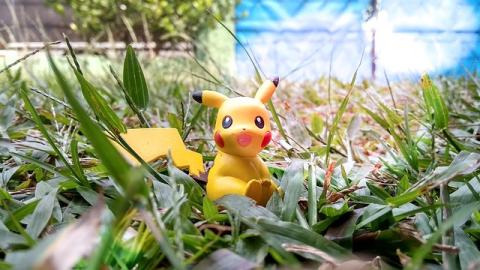 A happy Pikachu plastic model sitting in a green and grassy yard. There is a blue structure that is blurry in the background on the right side of the image.