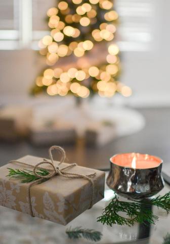 In the foreground, a wrapped gift tied with cord and decorated with a sprig of pine. Next to it, a candle in a small silver container burns with a pink glow. In the background is a lit and decorated tree with more gifts underneath it.