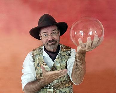 Professor Pathfinder holding a large bubble in his left hand at shoulder level. He is gesturing to the bubble with his right hand. He is wearing a button up vest with a mosaic of safari images on it, a white long sleeve button up shirt with the sleeves rolled up to his elbows, and a black fedora. Professor Pathfinder is standing in front of an orange background.
