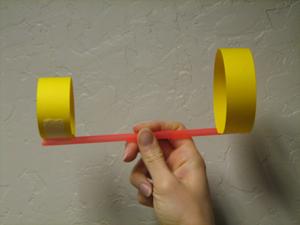 A hand holding a glider in the middle of its orange straw body. The wings are 2 yellow, hollow circles of paper that have been attached to either end of the straw. The circle on the left is smaller than the circle on the right. The glider is being held in front of a white wall.