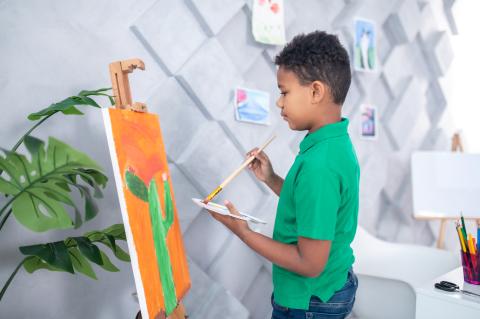 A young boy looking focused in a room with gray walls that have protruding square panels. The boy is painting a green cactus on an orange background. There are green plant leaves in the lower left hand corner.