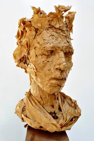 A bust of a person with spiky hair made from pieces of torn, brown butcher paper. The bust is looking forward at a 45 degree angle to the right. There is a shawl made from larger pieces of brown butcher paper wrapped around the bust where the shoulders would be. The bust is in front of a white background.