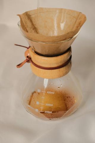 A homemade water filter made with a clear glass carafe and coffee filter placed in the top. There is a wood ring around the middle, thinnest part of the carafe, and a small amount of brown liquid pools in the bottom of the carafe.
