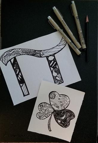 Example Zentangles. The image at the top is a pi symbol, and the image at the bottom is a three leaf clover. Both are full of zentangles drawn in black fine tipped pens. Three of these pens and a pencil are resting in the upper right hand corner.