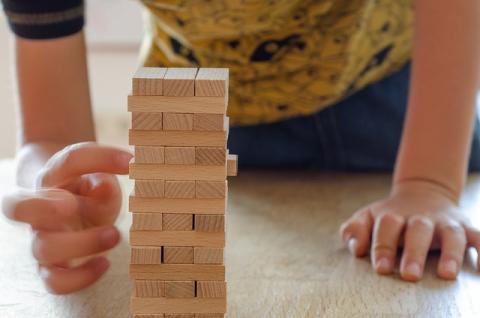 A young person plays a Jenga game. The stack of blocks is in the foreground and the person is on their hands and knees to play the game. They are in the process of pushing out the first block. 