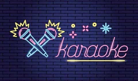 A cartoon neon karaoke sign on a navy brick wall. There are 2 cartoon neon microphones laying in an X shape on the left side of the wall. On the right side is the word “karaoke” in pink cartoon neon letters and underlined in yellow. 