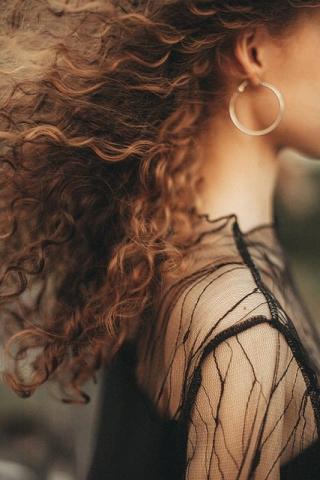 A photograph of a woman standing in profile to the camera. You cannot see her face, but instead focus on her brown, curly hair that is being lifted behind her by a breeze. She is wearing a black dress with black mesh long sleeves.