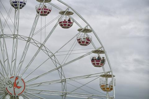 A ferris wheel in front of a gray, cloudy sky. The frame of the ferris wheel is white, and it has red cars to hold the people.