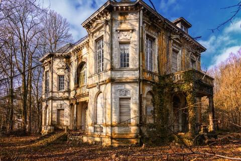 A haunted looking chateau on an abandoned looking lot of land. The building is old and dirty with ivy clinging to some places. There are trees in the background with no leaves on them. The ground seems to be covered in dead greenery. 