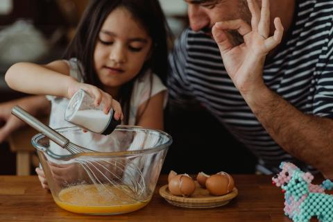 At a kitchen table, a pre-teen girl adds salt to a bowl of beaten eggs. Her father stands next to her indicating to add "just a pinch." 