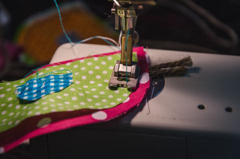 A stack of cut fabric pieces – blue gingham, small polka dots on green and large polka dots on red  - sit under the presser foot of a sewing machine.