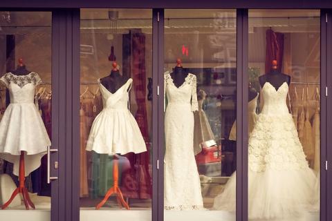 A storefront window with 4 white wedding dresses on black mannequins. The 2 dresses on the left are shorter and would end at the knee. The 2 dresses on the right reach the floor. 