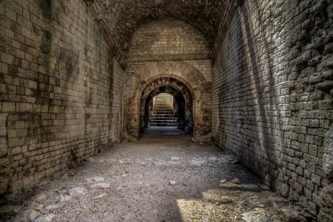 A large, empty corridor with stone walls and floor. There is a staircase leading up and out in the back of the picture.