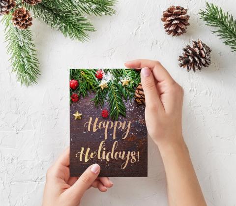 Person holding a seasonal greeting card with both hands. The card says Happy holidays along with an image of pinecones and fir. Behind the image there is an adornment with pine cones and fir.   Persona que sostiene una tarjeta de felicitación de temporada con ambas manos. La tarjeta dice Feliz fiestas junto con una imagen de piñas y abetos. Detrás de la imagen hay un adorno con piñas y abeto.