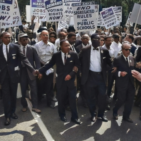 A colorized photo of Martin Luther King Jr. walking in protest with other men in suits.