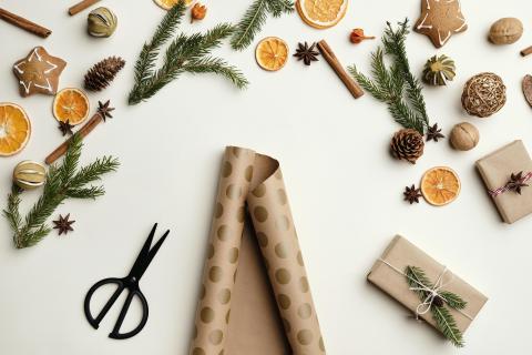 pair of scissors and tan wrapping paper with gold dots on a white surface, surrounded with pine sprigs, holiday cookies, pinecones, orange slices, cinnamon sticks, star anise and two small wrapped presents