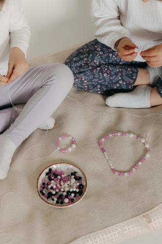 two children sitting on floor placing beads on a string to form a necklace